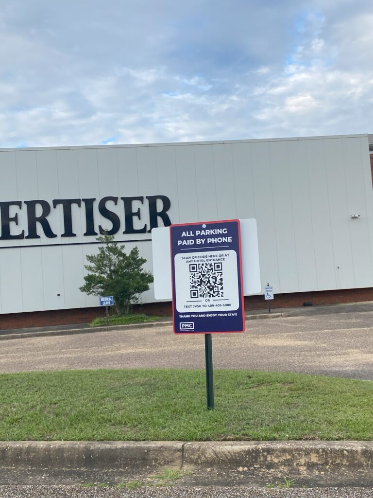 A sign in a parking lot shows instructions for phone payment, including QR codes and a number to text. A large building with partial signage is in the background, along with grass, pavement, and a few trees.