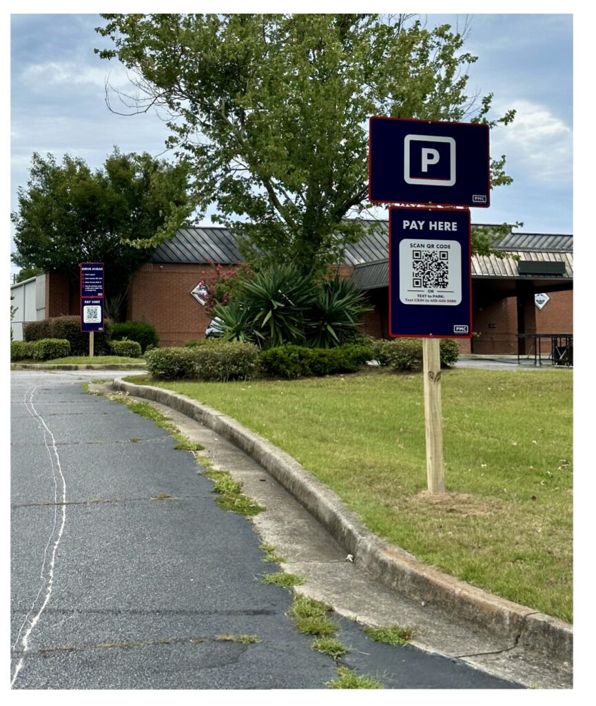 A parking lot with a "P" parking sign and a "Pay Here" sign featuring a QR code. A brick building and trees are in the background, with grass and a curb in the foreground.