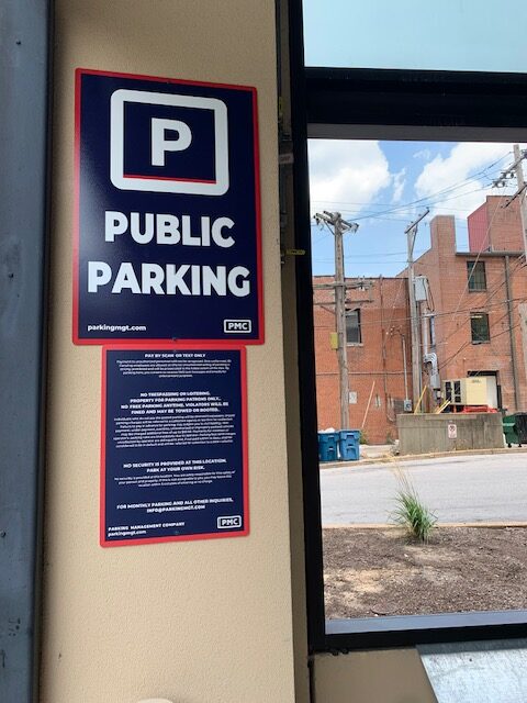 A sign on a wall reads "Public Parking" in bold letters, with a large white PMC logo in a blue square above it. Below is additional information. Outside the window, brick buildings, power lines, and a dumpster are visible.