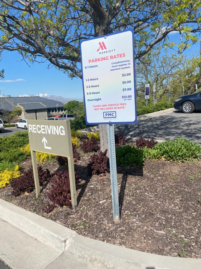 A sign near a Marriott hotel parking lot lists parking rates and durations, with a “RECEIVING” sign and arrow beside it. Trees, bushes, a road, and distant mountains are visible under a sunny sky.