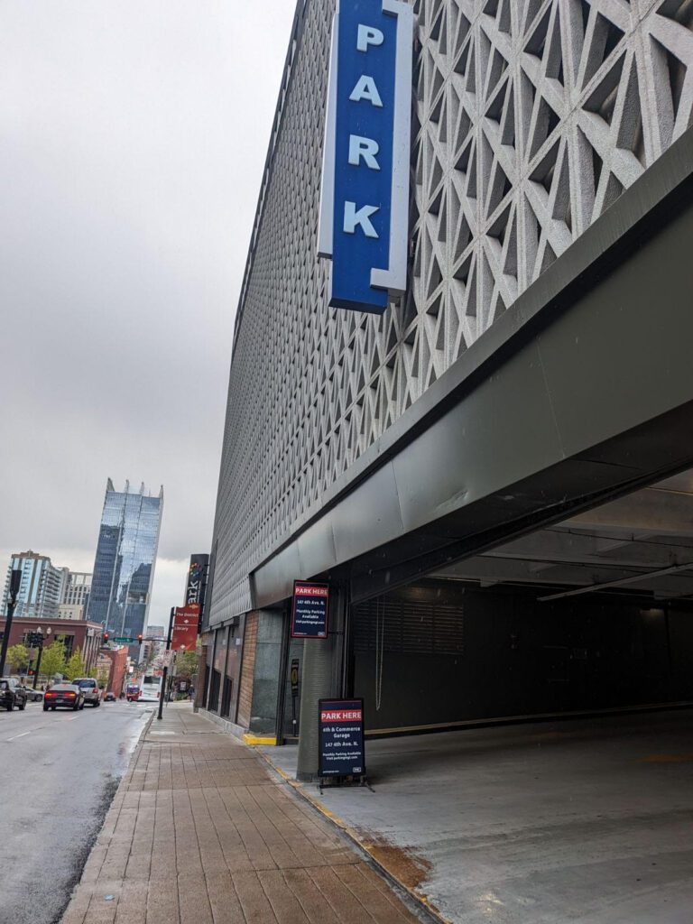 A city parking garage with a large blue "PARK" sign hangs above the entrance. The street is wet, and tall buildings, including one with a glass facade, are visible in the background under a cloudy sky.