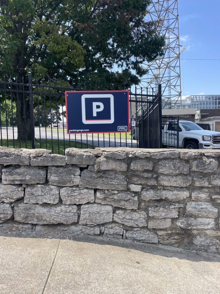 A parking sign mounted on a black fence behind a stone wall, with a white pickup truck and trees visible in the background under a sunny sky.