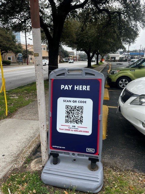 A parking sign on a sidewalk reads "PAY HERE" with a QR code for payment and a phone number to text. Cars are parked nearby with trees and buildings in the background.