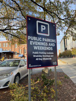 A sign reads "Public Parking Evenings and Weekends. Public Parking Available Weekday Evenings and All Day Weekends" on a city street with cars and buildings in the background.