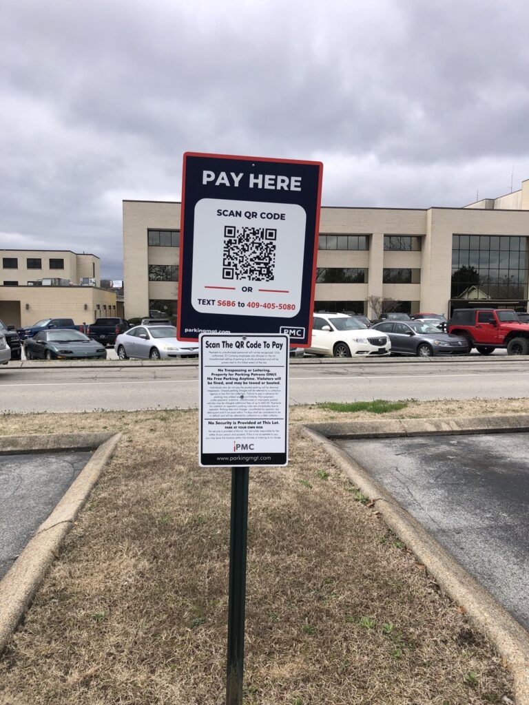 A parking lot sign reads “PAY HERE” with a QR code for payment. Below, instructions mention texting a number or scanning the QR code. Cars are parked and a large building is visible in the background under cloudy skies.
