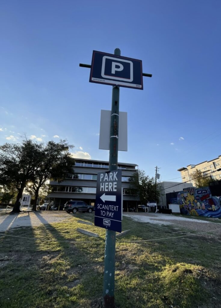 A parking sign stands in front of an outdoor parking lot, with a building and a mural visible in the background. The sign reads "PARK HERE" and "SCAN/TEXT TO PAY" under a large "P" for parking.