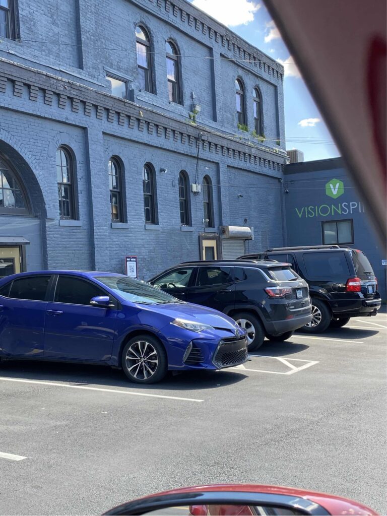 Three cars are parked in a lot beside a blue-gray brick building with arched windows. The building next door has a sign reading "VISIONSPIRE" and a blue sky with clouds is visible above.