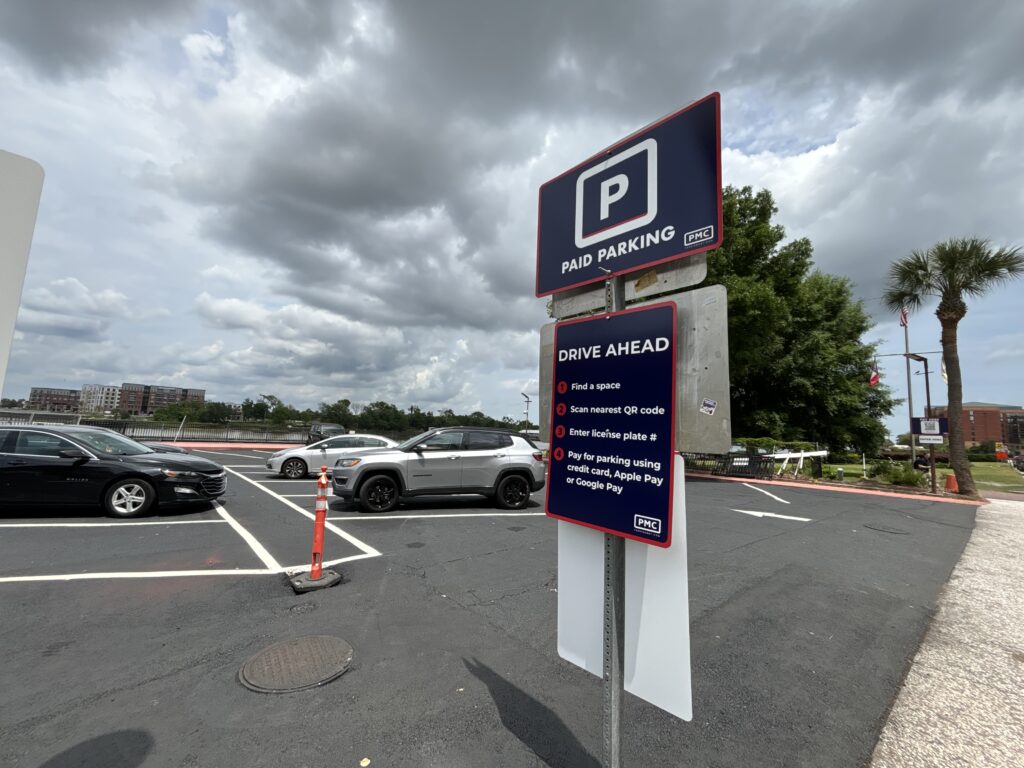 A paid parking lot under a cloudy sky with a sign listing parking instructions, several parked cars, and trees in the background.