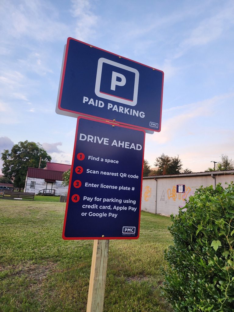 A blue and red PMC sign displays "Paid Parking" and instructions: find a space, scan QR code, enter license plate, and pay by credit card, Apple Pay, or Google Pay. The sign stands on grass near a building.