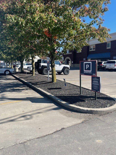 A parking lot with several cars parked near a building. Trees with green leaves line the edge of the lot, and signs indicate public parking rules. The weather is sunny and clear.