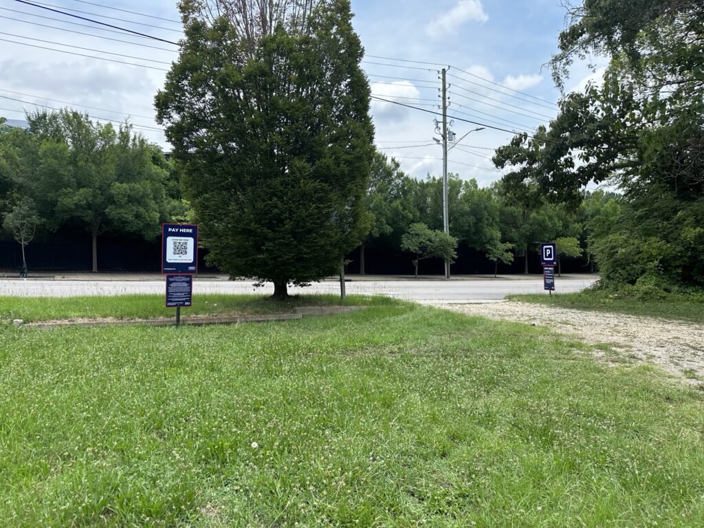 A grassy area with two parking signs near a road, one sign displaying a QR code for payment. Trees and power lines are visible in the background under a partly cloudy sky.