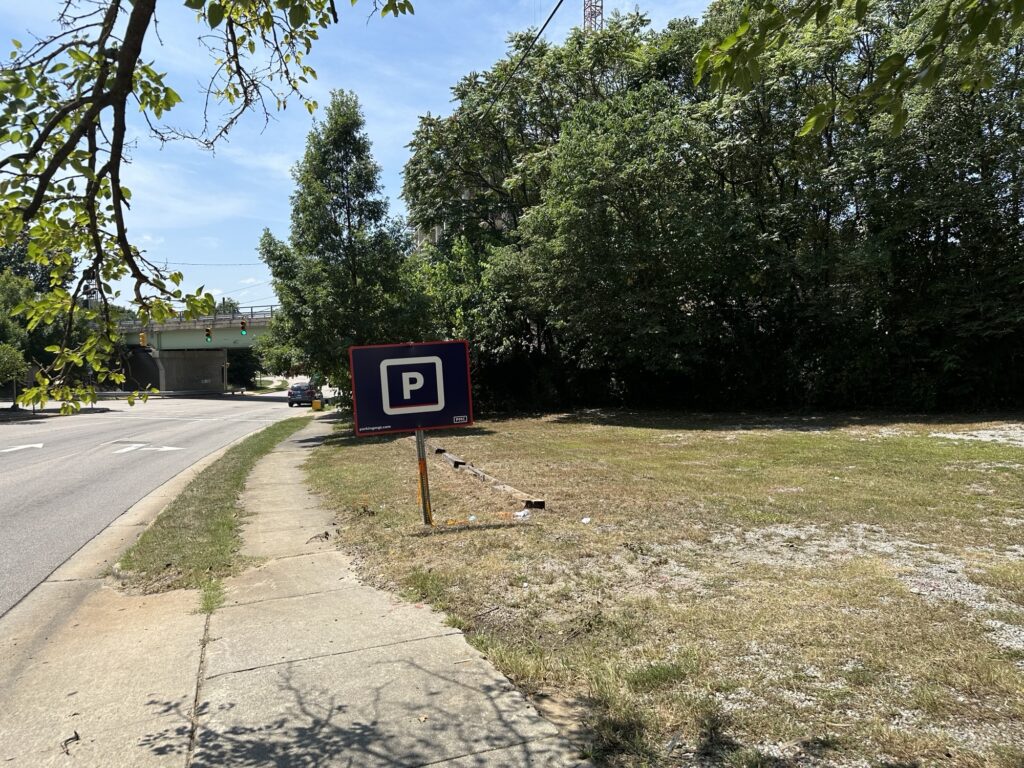 A PMC parking sign stands on a grassy lot beside a sidewalk and a road, with trees and an overpass visible in the background on a sunny day.