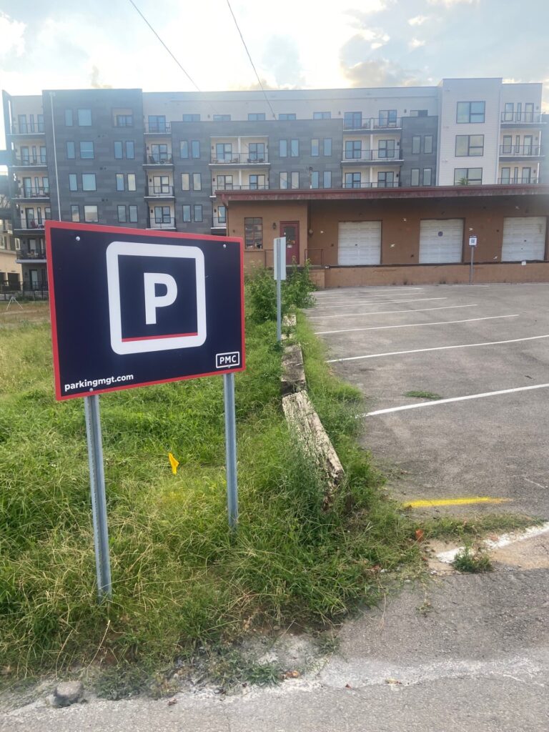 A parking lot with marked spaces and a large “P” parking sign in the foreground, set beside grassy overgrowth, in front of a modern multi-story apartment building.