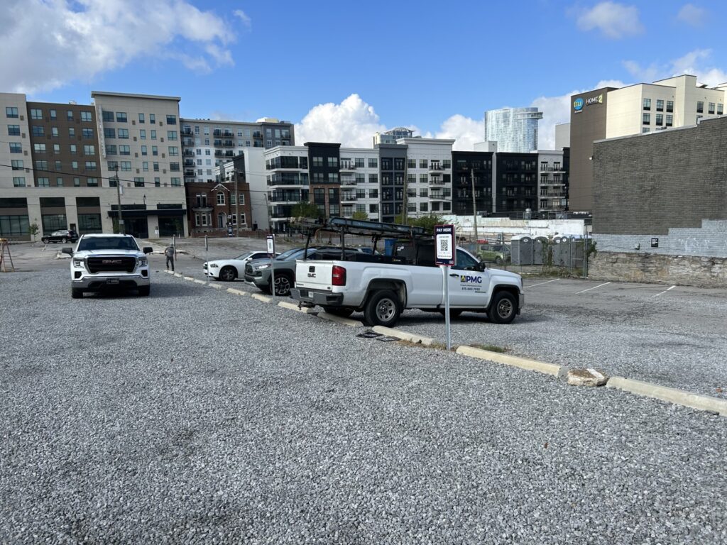 A mostly empty gravel parking lot with a few parked vehicles, including a white pickup truck with a ladder on top. Modern apartment buildings and a blue sky with clouds are visible in the background.