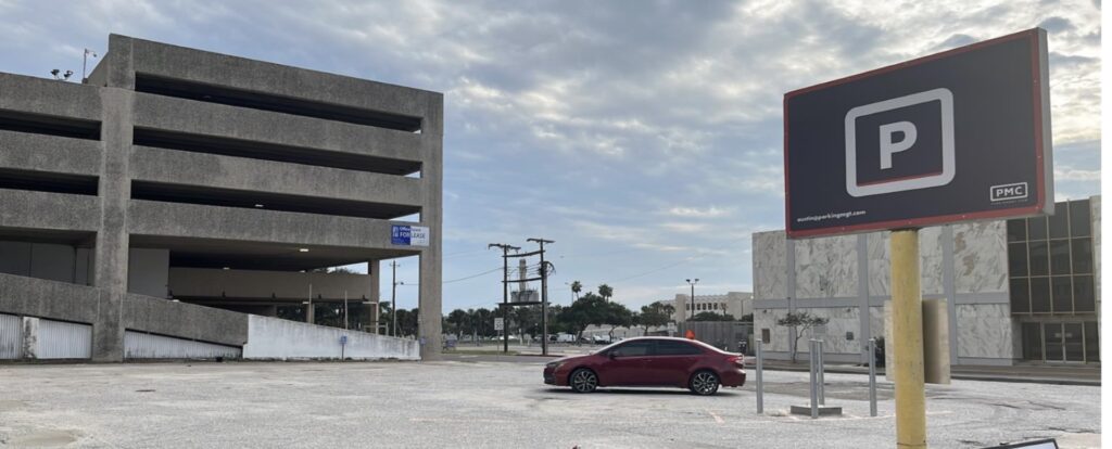 A nearly empty parking lot with a single red car parked near a concrete parking garage. A large sign with a white "P" on a black background indicates parking. The sky is partly cloudy.