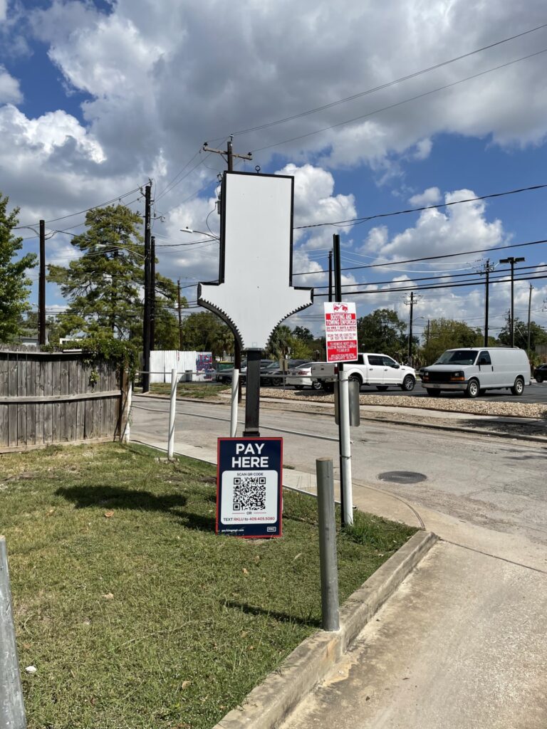 A large, blank arrow-shaped sign stands by a roadside parking lot. Below it is a "Pay Here" sign with a QR code. Cars and utility poles are visible under a partly cloudy sky.