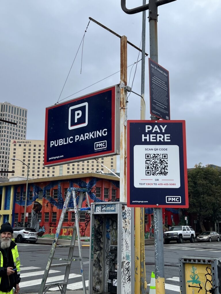 Two signs on a city street show information for public parking, including a QR code for payment and instructions to pay by text. A man stands on the left, with colorful buildings and tall city structures in the background.