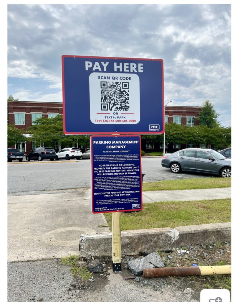 A parking lot with a large blue sign that says “PAY HERE” above a QR code for payment. An additional sign below lists parking management rules. Several cars and a building are visible in the background.