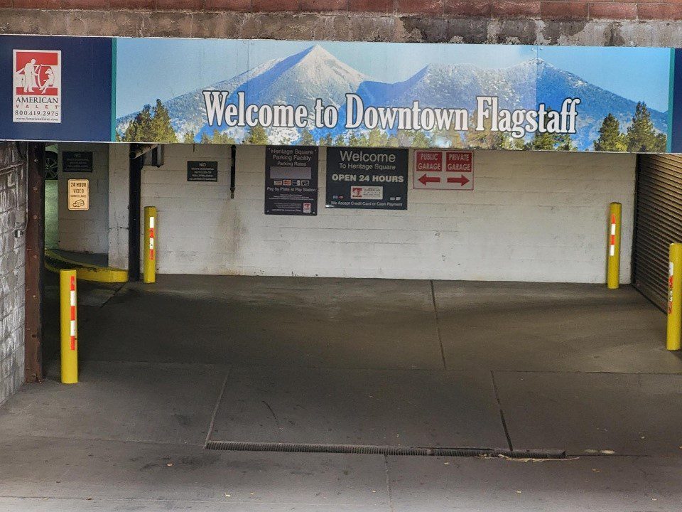 Entrance to a parking garage with a sign above reading "Welcome to Downtown Flagstaff" and an image of mountains and trees in the background. There are yellow poles and additional directional signs in the garage.