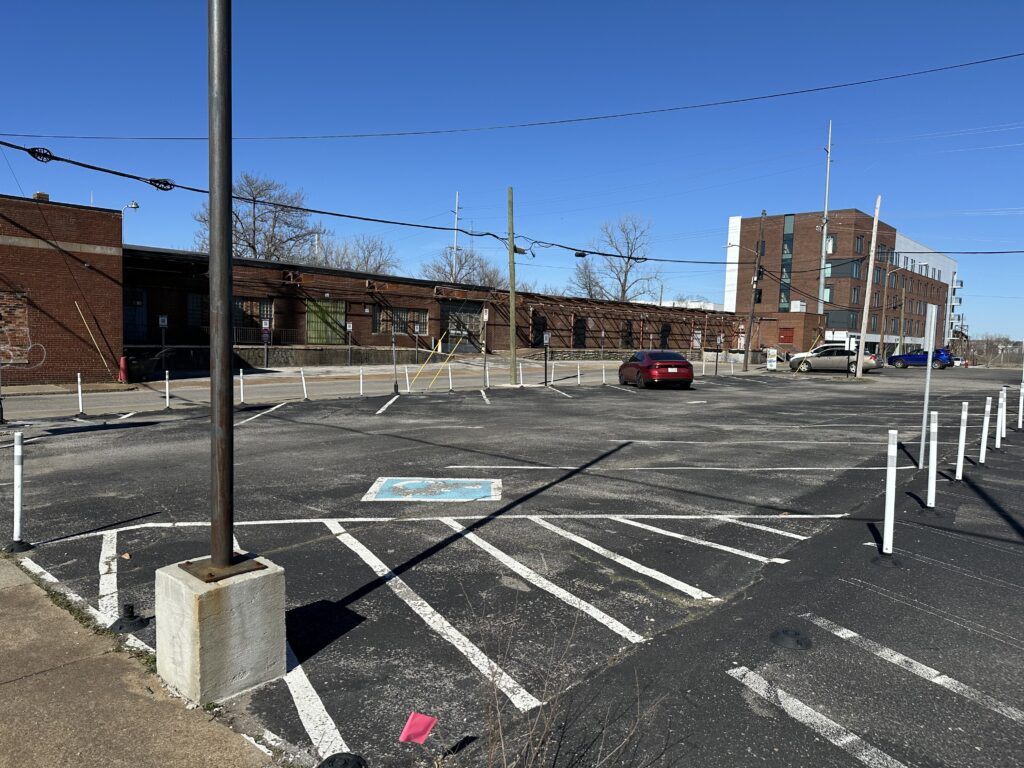 An almost empty parking lot with faded white lines, a single parked red car, a building in the background, and a blue handicap parking space in the foreground, all under a clear blue sky.