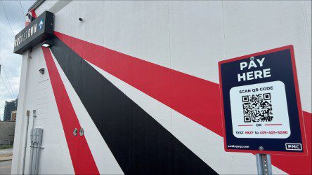 A sign on a white wall with red and black stripes says "Pay Here" and displays a QR code for payment. The building has bold diagonal lines and a sign reading "POST HTX" near the roof.