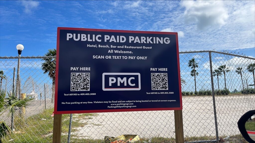 A large sign reading "Public Paid Parking" with QR codes for payment stands in front of an empty lot. A chain-link fence, palm trees, and a partly cloudy blue sky are in the background.