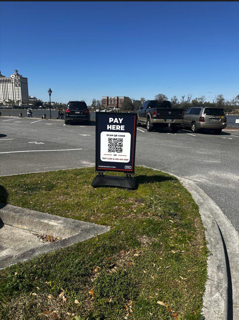 A PMC sign in a parking lot reads "PAY HERE" with a QR code for payment. Several parked cars and buildings are visible under a clear blue sky in the background. Grass and pavement are in the foreground.