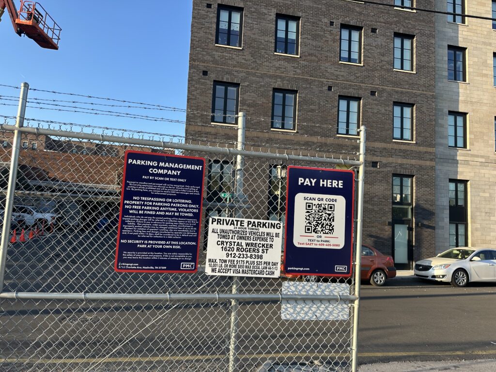 A fenced Parking Management Company parking lot with PMC signs about parking rules and payment instructions. A QR code for payment is visible. An apartment building and parked cars are in the background.
