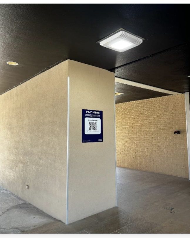 A beige wall with a black ceiling above features a sign that reads "PAY HERE" next to a QR code, located in a mostly empty, covered walkway area.