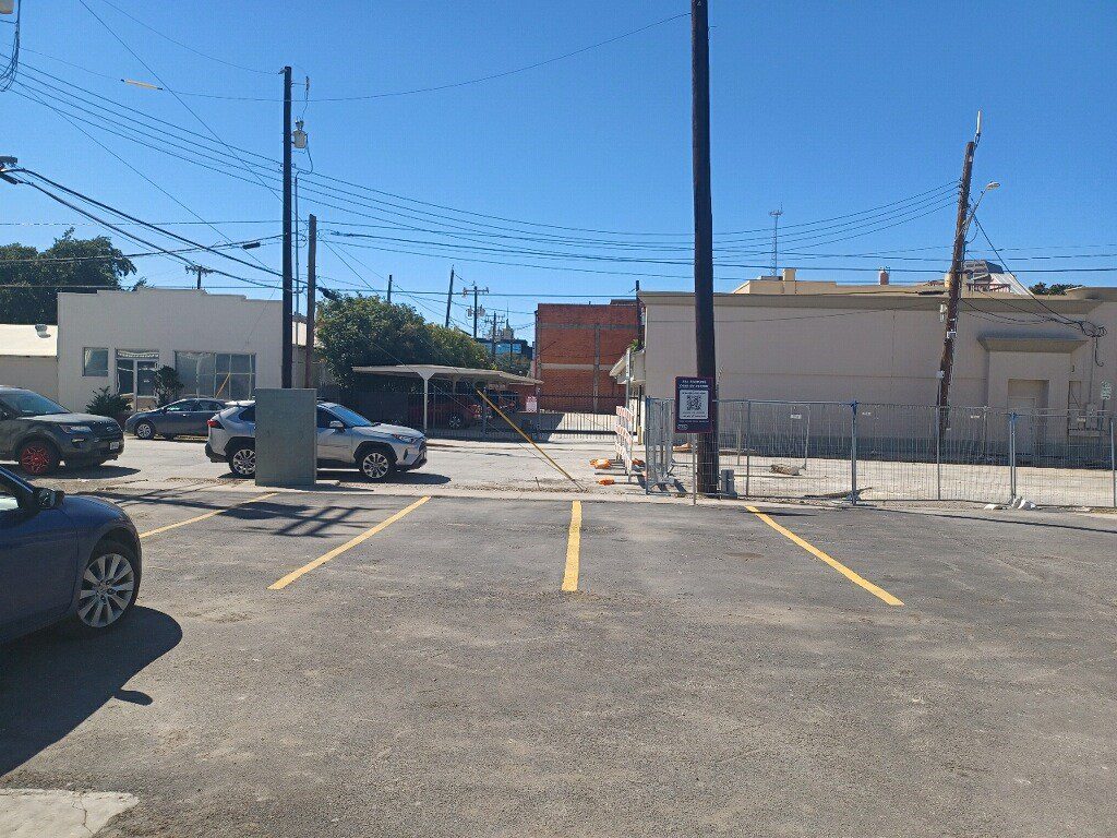 A mostly empty parking lot with a few parked cars, yellow-painted parking lines, utility poles, and some buildings in the background under a clear blue sky.