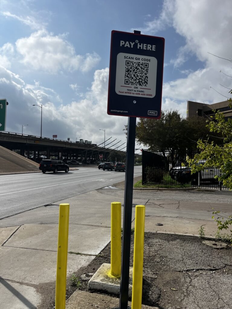 A "Pay Here" parking sign with a QR code stands on a sidewalk next to a street with cars. Yellow posts protect the sign, and a highway overpass is visible in the background under a partly cloudy sky.