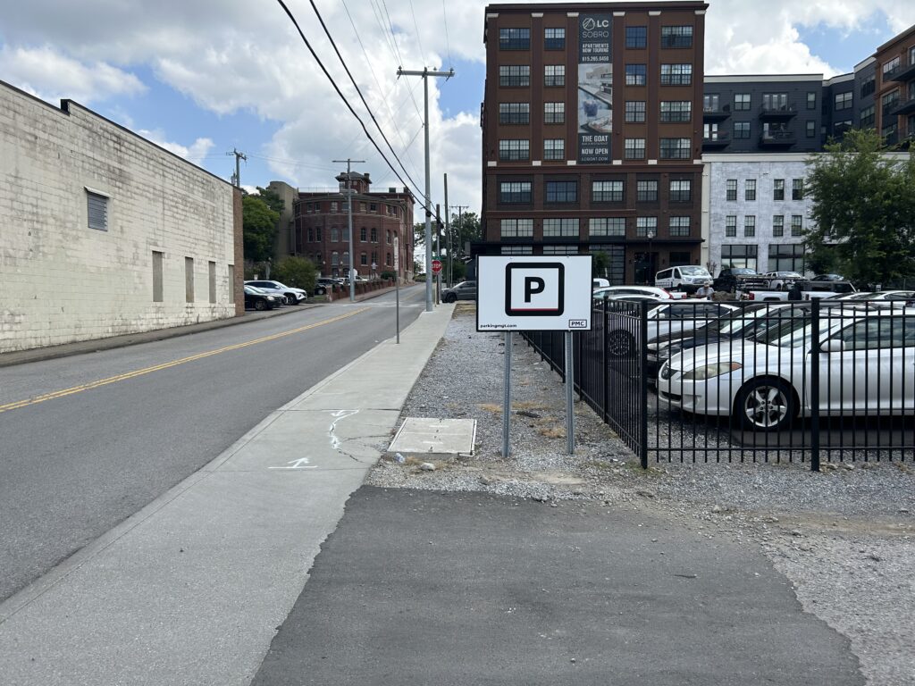 A sidewalk runs beside a street with a visible "Parking" sign next to a fenced parking lot filled with cars. Brick and white buildings are in the background under a partly cloudy sky.