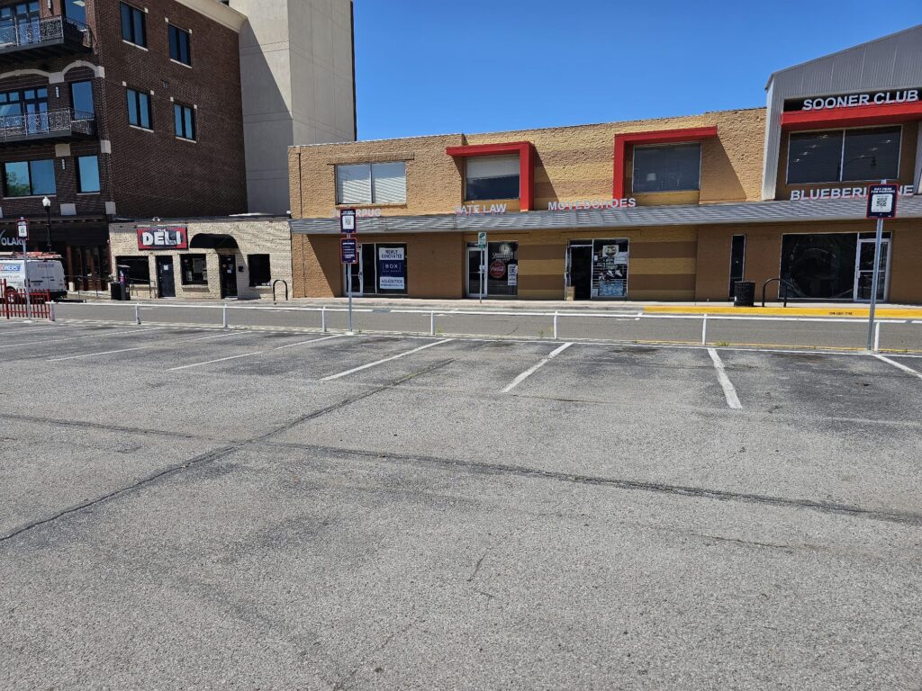 A mostly empty parking lot in front of a row of businesses, including a deli and a store with a "Gateway" sign in red letters. The buildings have brick and tan facades, with several large windows and doors.