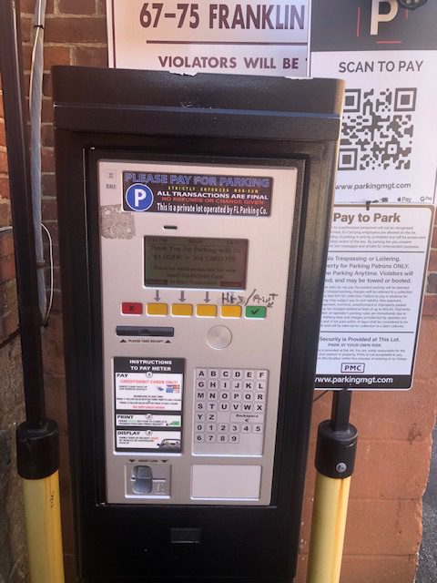 A parking payment machine mounted on a wall with instructions, a keypad, card slots, and a digital screen. A sign above displays an address, QR code, and additional payment information.