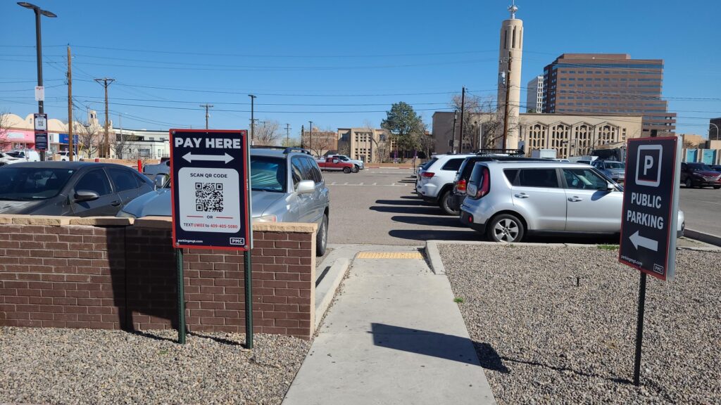A parking lot with several cars parked. Signs indicate “Pay Here” with a QR code and “Public Parking” with an arrow. A church and tall office building are visible in the background under a clear blue sky.