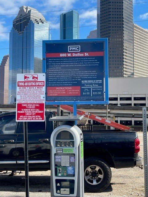 Parking payment kiosk and information signs in a downtown Houston parking lot, with tall skyscrapers in the background and a pickup truck parked nearby under a sunny sky.