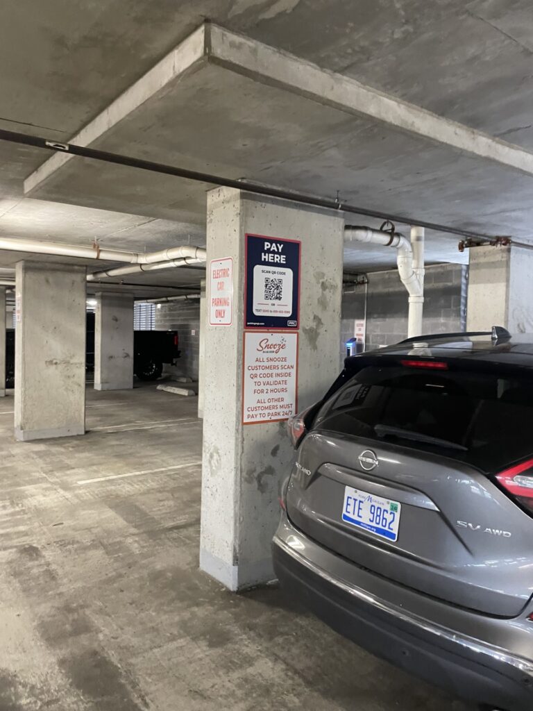 A gray Nissan SUV parked in an indoor parking garage next to a pillar with a sign displaying parking payment instructions and a QR code. The area is lit by natural and artificial light.