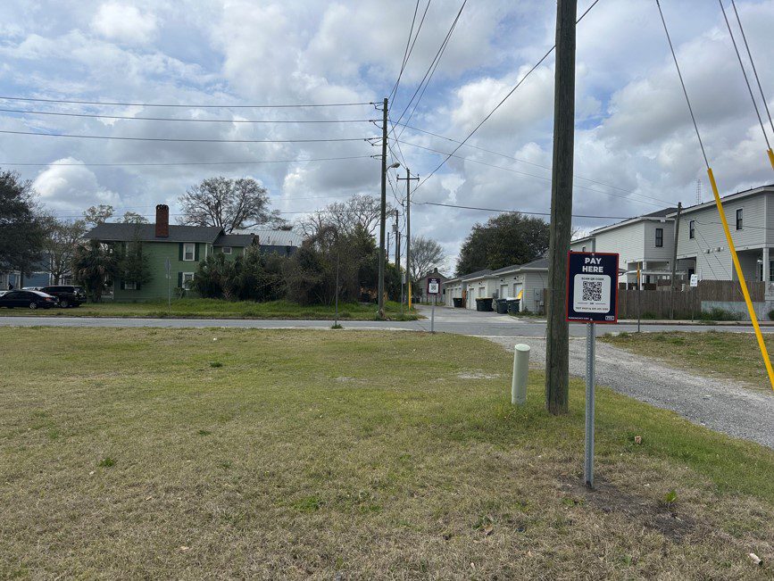 A grassy lot with a Parking Management Company "Pay Here" parking sign featuring a QR code for payment. A gravel road runs along the right side of the image.