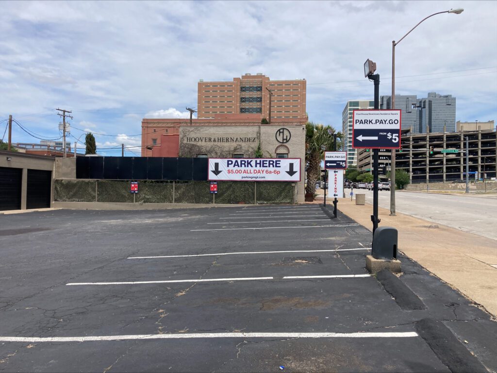 An empty parking lot with signs displaying “PARK HERE $5.00 ALL DAY 6-9” and “PARK-PAY-GO $5.” A tall building and a parking garage are visible in the background under a blue sky.