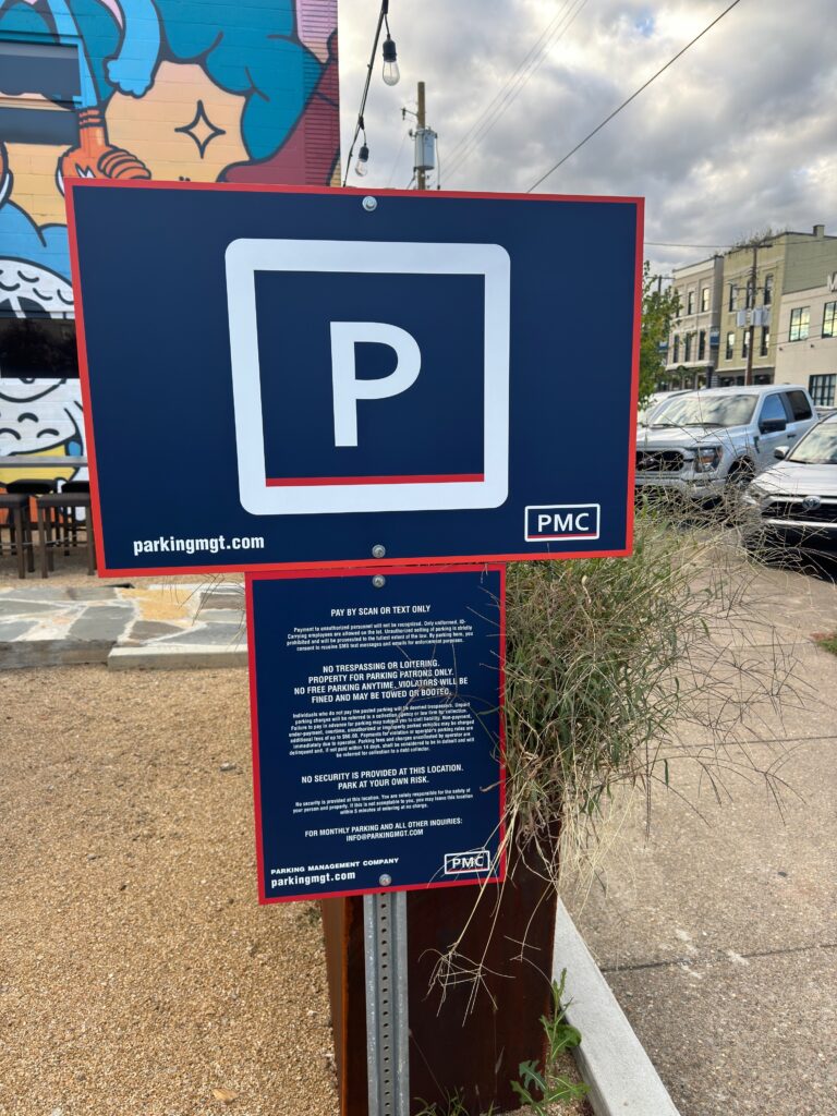 A blue parking sign with a white "P" and information below stands near a street with parked cars, a mural, and a building in the background under a partly cloudy sky.