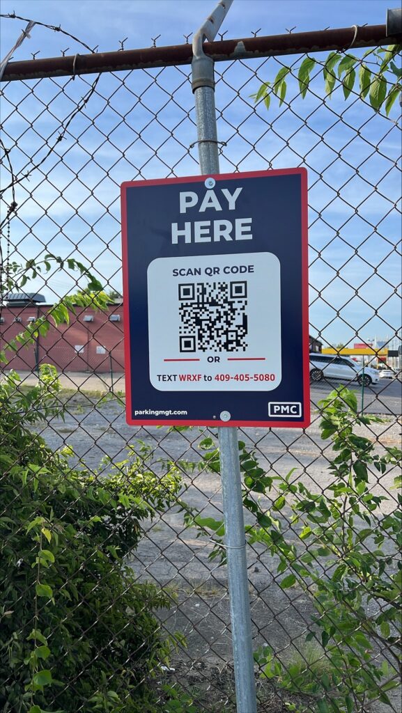 A sign attached to a chain-link fence reads "PAY HERE" with a QR code and instructions to scan or text WRXF to 409-405-5080 for payment. Bushes and a building are visible in the background.