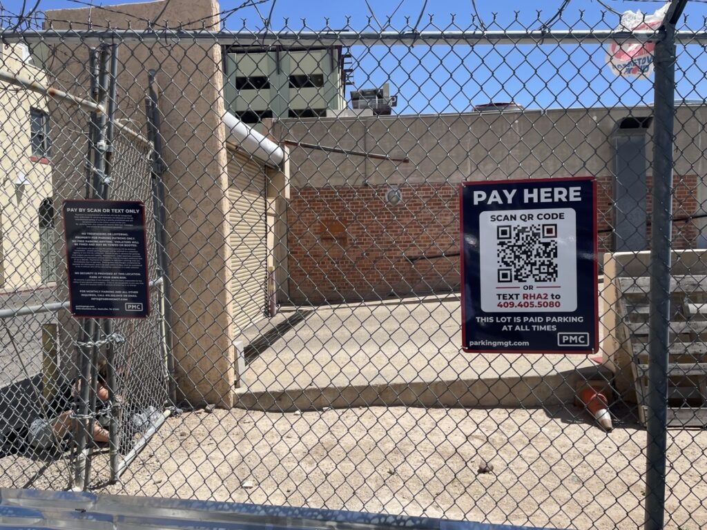 A fenced parking lot with a PMC "Pay Here" sign featuring a QR code for payment and phone number for customer support. Another sign with parking instructions is posted on the left side of the fence.