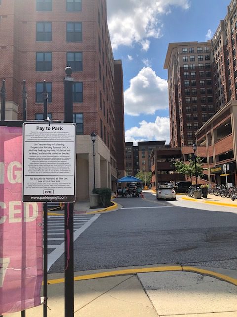 A city street lined with tall brick buildings features a "Pay to Park" sign in the foreground and a blue sky with scattered clouds overhead. A few bicycles and a tent are visible further down the street.
