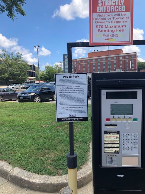 A parking payment machine stands near a "Pay to Park" sign and another sign warning of a $70 maximum booting fee. Cars are parked in the background, with trees, grass, and a building visible under a partly cloudy sky.