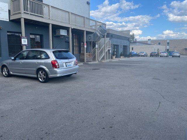 A silver hatchback car is parked in a mostly empty asphalt parking lot beside a gray building with a wooden balcony. Several other cars are parked in the distance under a blue sky with scattered clouds.