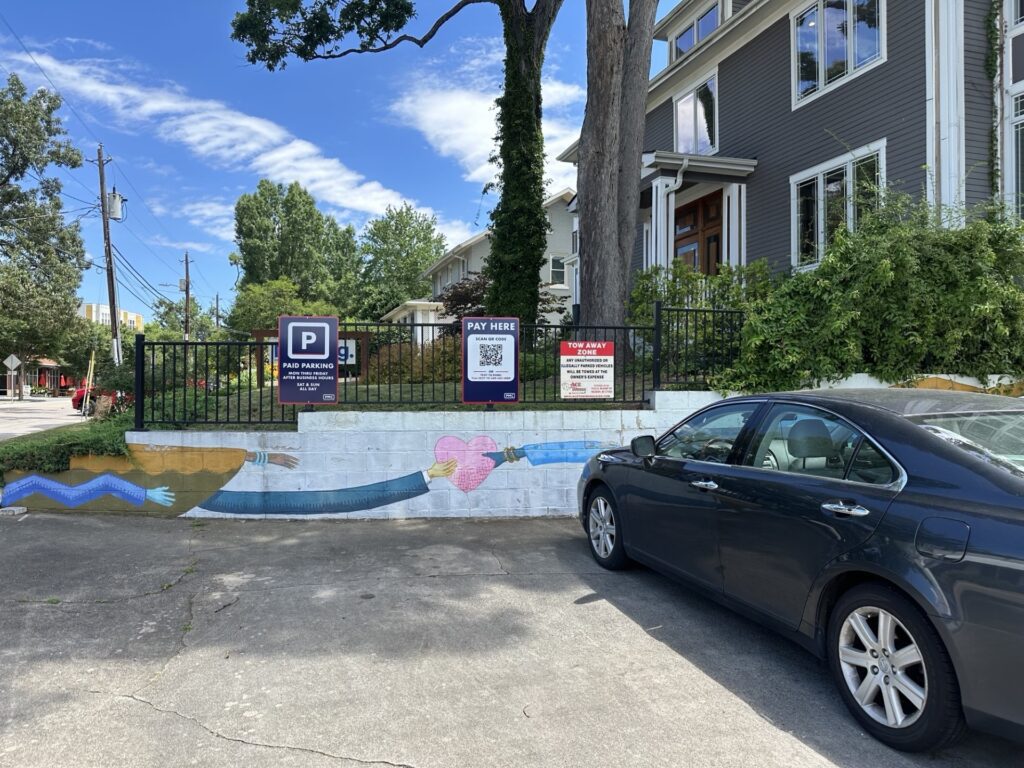 A black car is parked near a painted mural of two arms holding a heart on a low wall. Nearby are PMC parking signs and a "Pay Here" station with a QR code for digital payment. A gray house and trees are in the background.