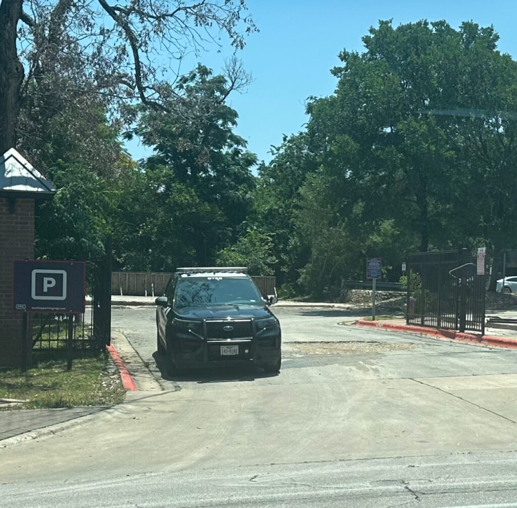 A black police SUV is parked at the entrance of a gated lot on a sunny day, with trees and a "P" parking sign visible nearby.