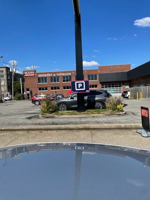 Cars are parked in a lot near a brick building labeled "AKASHA Brewery & Taproom." A parking sign is mounted on a post in the center, and the sky is clear and blue.