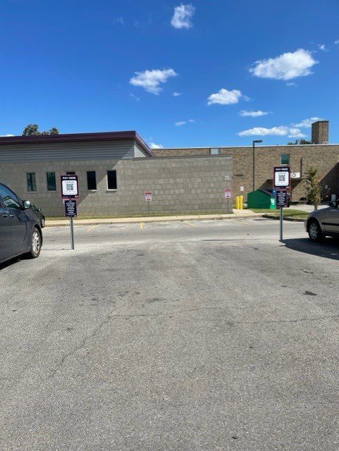 A mostly empty parking lot on a sunny day, with two cars parked on either side. Signs in the center indicate reserved or special parking spaces. A brick building and blue sky with clouds are in the background.