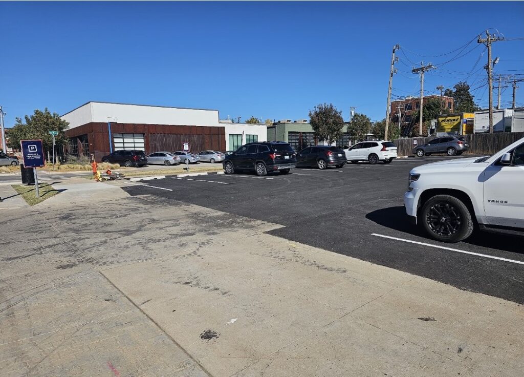 A PMC-owned parking lot with a few parked cars on a sunny day. A modern building and some trees are in the background, with utility poles and wires visible on the right side.